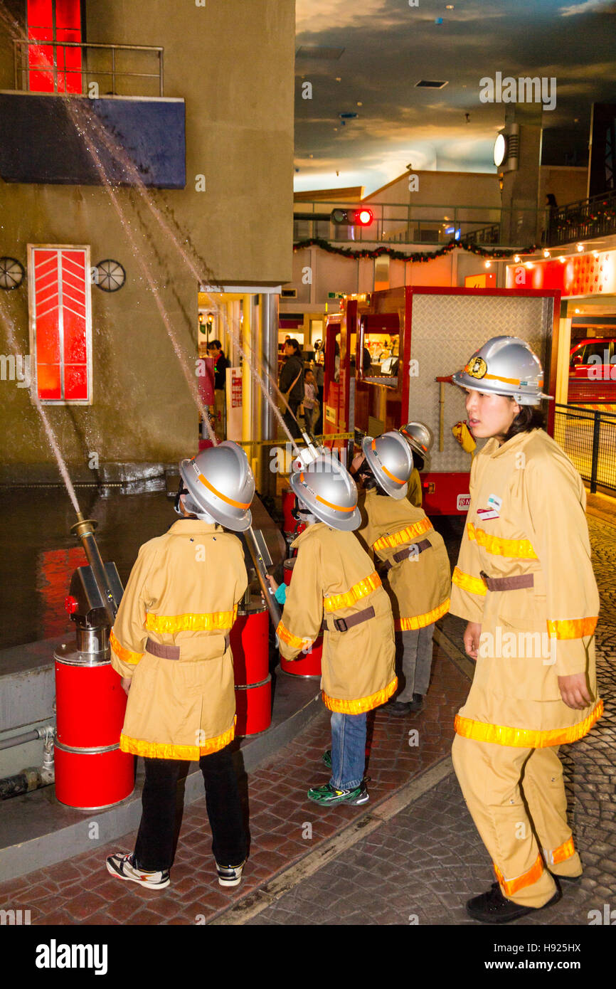 Giappone, Nishinomiya, KidZania, attività figlio il centro del gioco. Linea di bambini vestiti come i vigili del fuoco mirando tubi flessibili a combustione edificio. Foto Stock