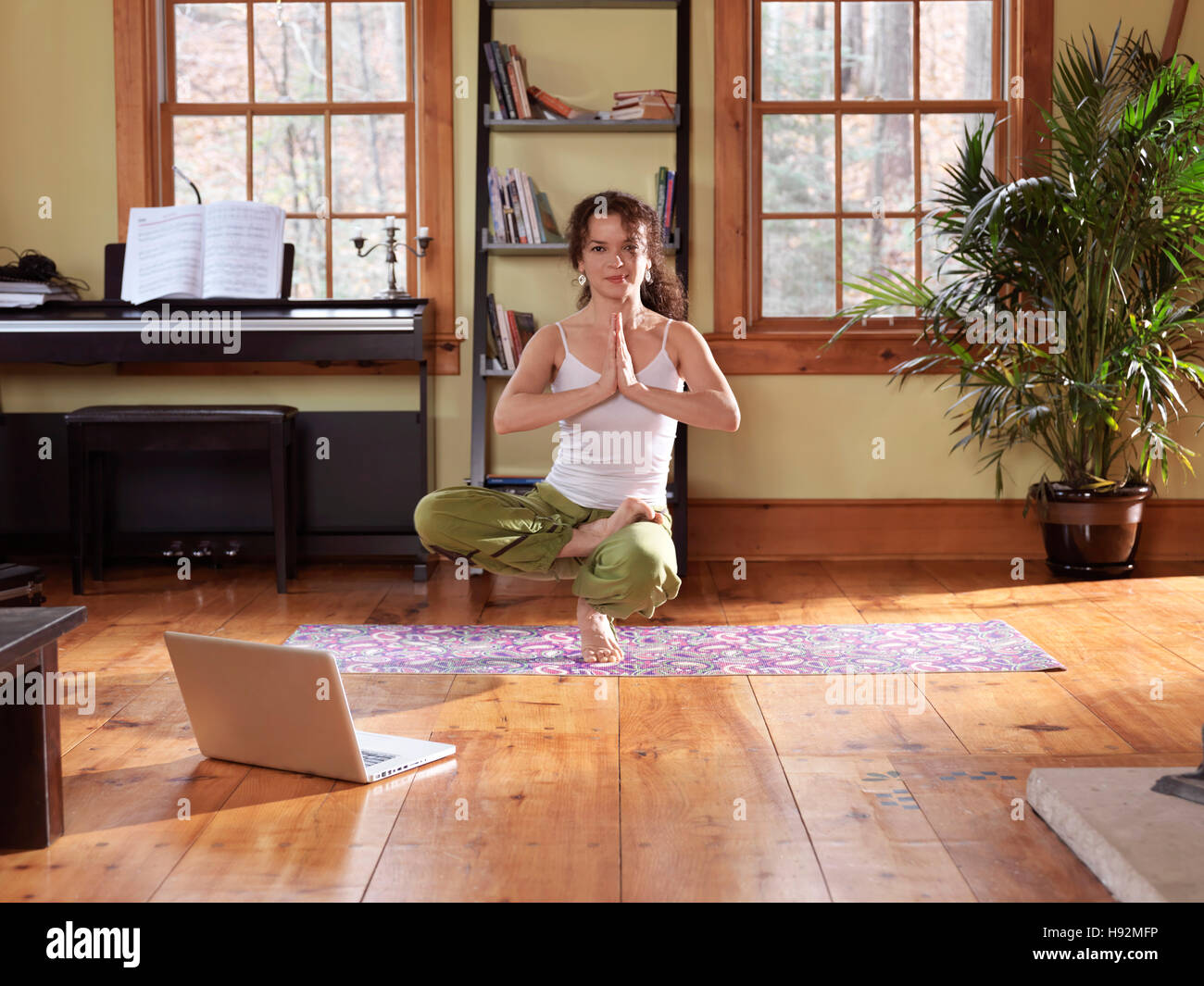 Giovane donna a praticare yoga, in equilibrio su un piede a casa in un salotto con un computer portatile dal suo lato Foto Stock
