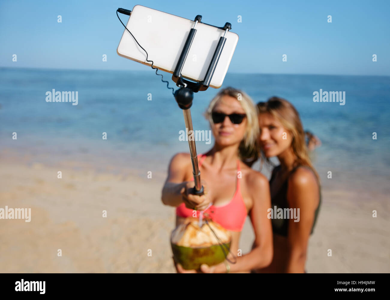 Due giovani amici di sesso femminile sulla spiaggia prendendo autoritratto con telefono cellulare su selfie stick. Le donne in vacanza sulla spiaggia prendendo selfie. Focus su smart pho Foto Stock