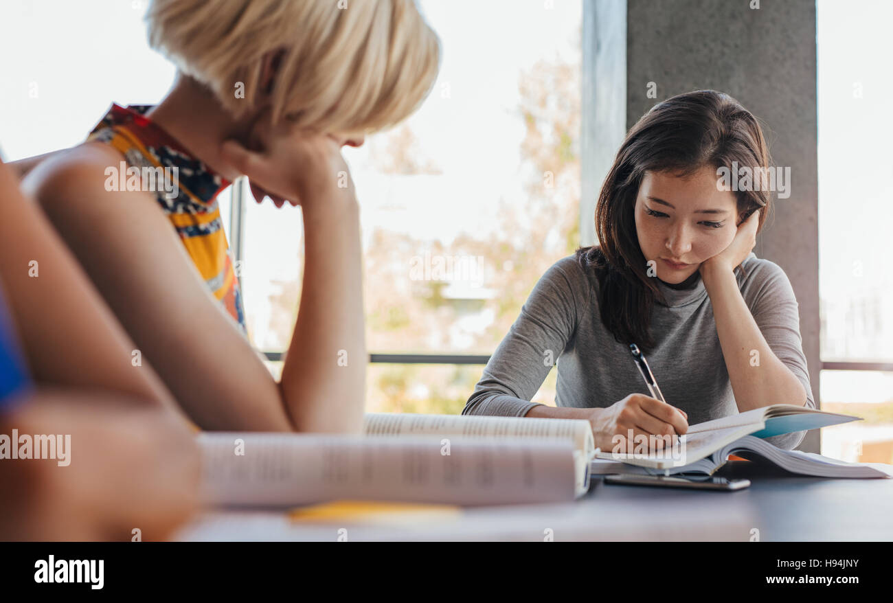 Giovane donna asiatica la scrittura su notebook con i compagni di classe lo studio intorno nella biblioteca universitaria. Gli studenti preparare il disco per gli esami finali. Foto Stock