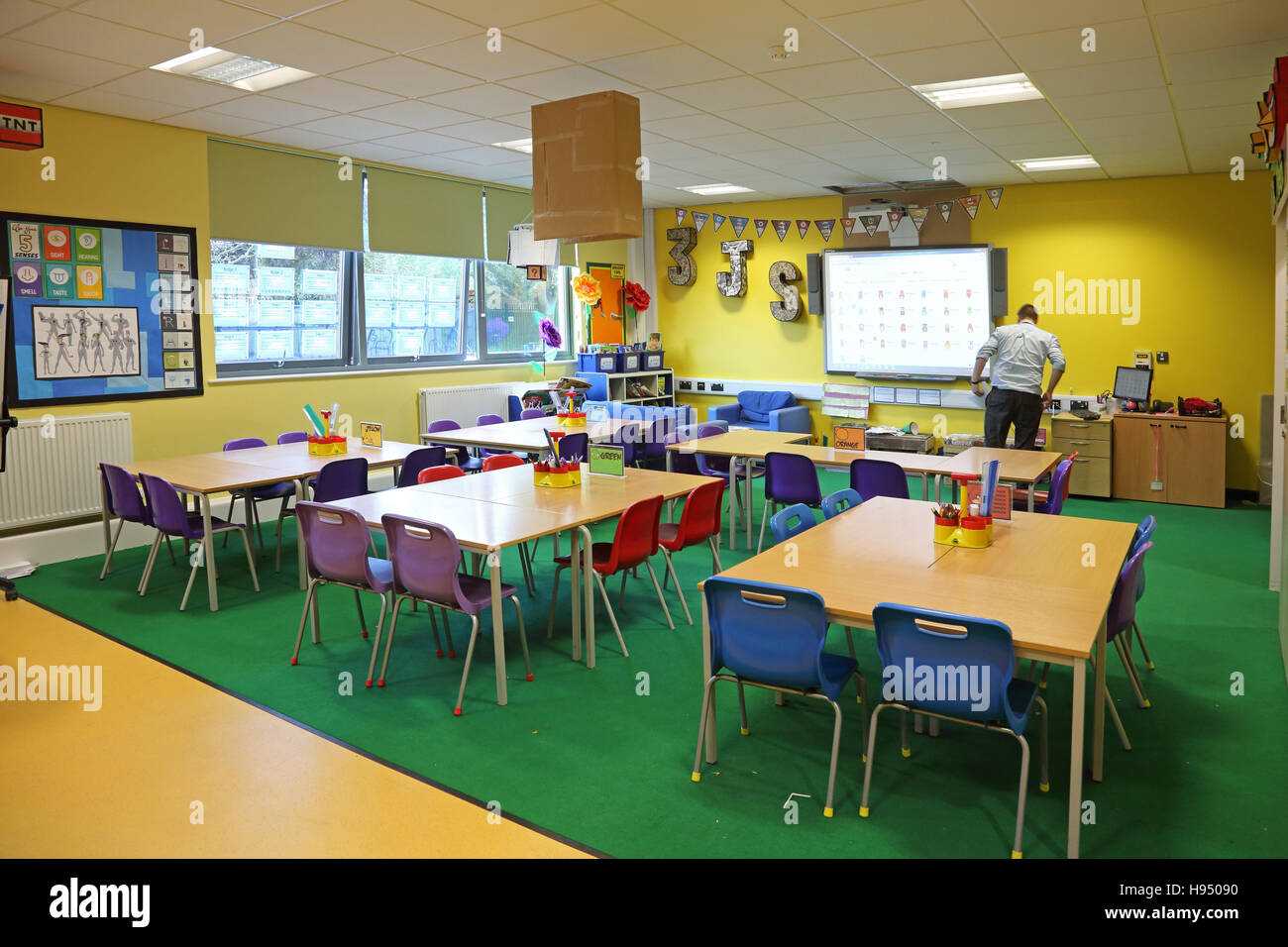 Vivacemente colorato interno di un moderno anno 1 aula scolastica che mostra tavoli e sedie e lavori artistici sulle pareti. Mostra insegnante. Foto Stock
