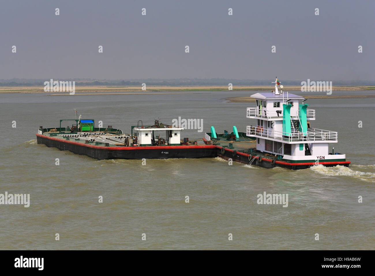 Un fiume in barca da carico capi a monte spingendo una chiatta caricato con tubo in calcestruzzo sul fiume Irrawaddy in Myanmar (Birmania). Foto Stock