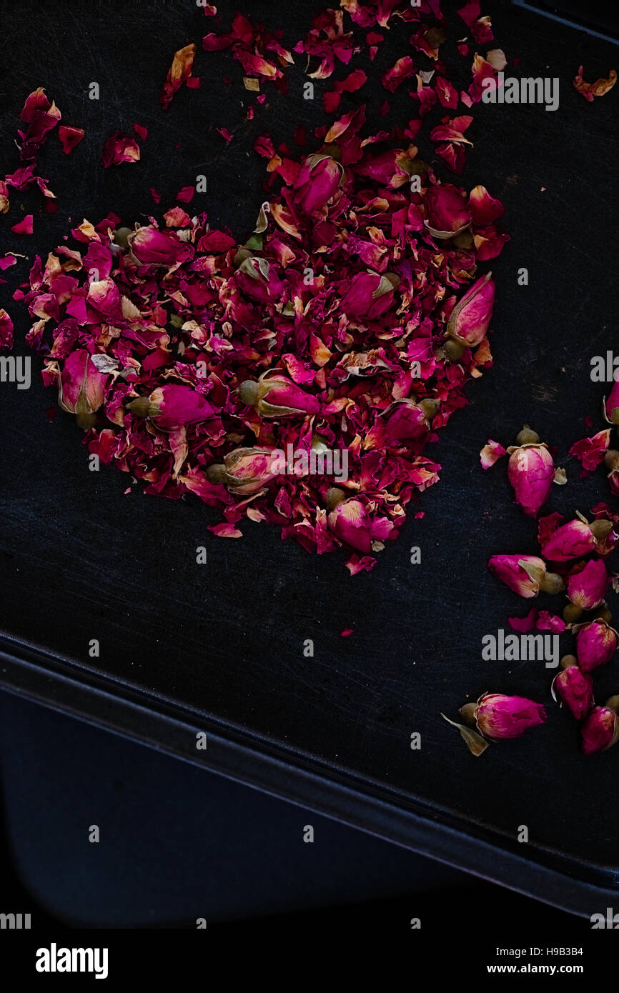 Asciugato i petali di rose e gemme in una forma di cuore su uno sfondo scuro. Ingrediente romantico per il tè, vasca da bagno o cosmetici. Copia dello spazio. Dark cibo fotografia Foto Stock