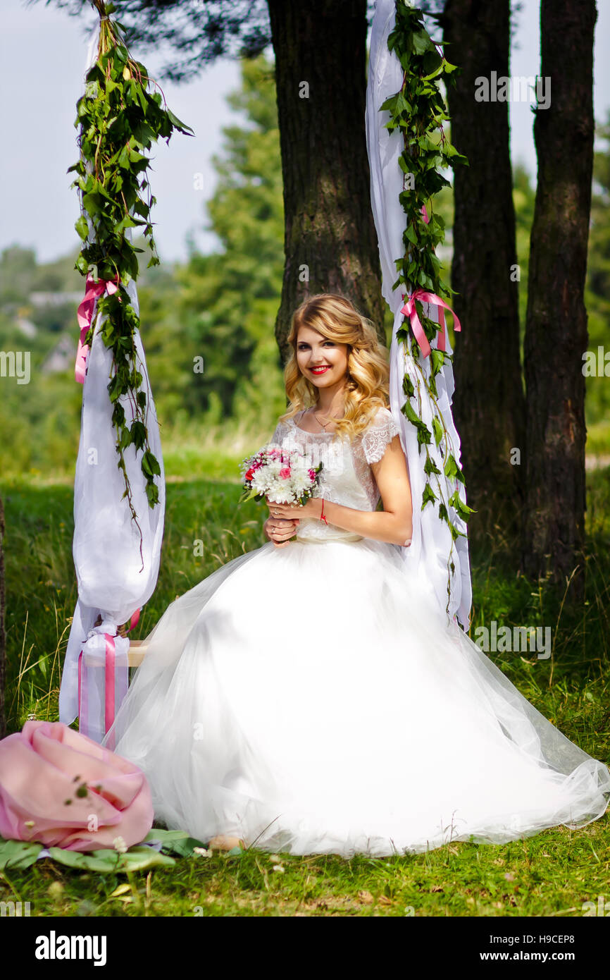 Bella bionda sposa in un bianco abito da sposa oscilla su decorate wedding swing e sorridente alla telecamera, estate, bouquet Foto Stock