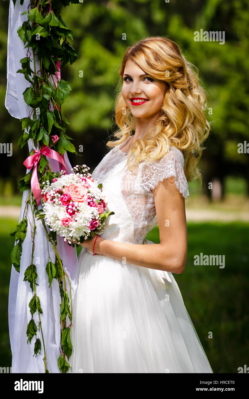 Bella bionda sposa in bianco di un abito da sposa in piedi nella parte anteriore del decorate wedding swing, estate, bouquet di nozze Foto Stock