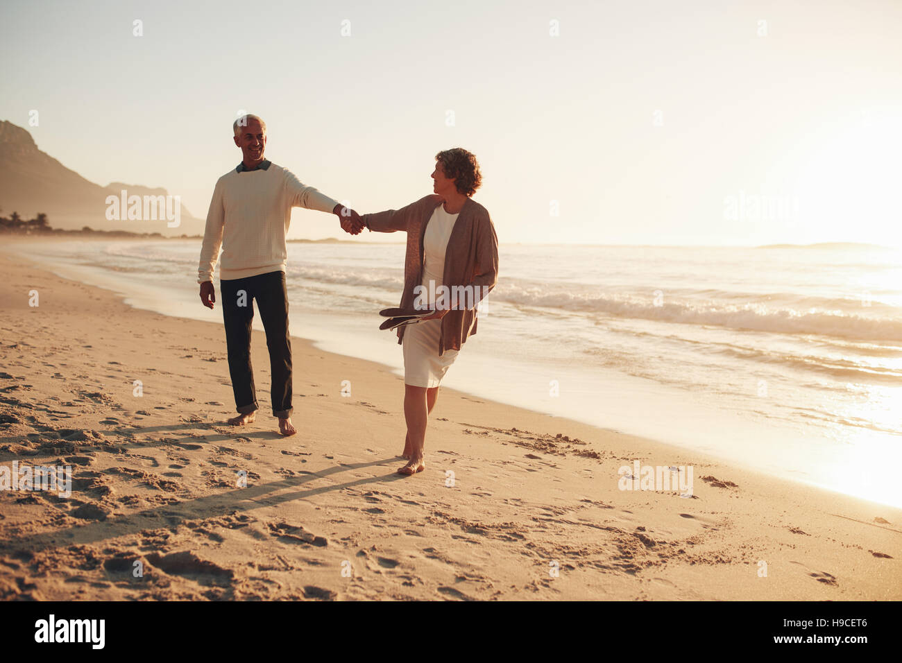 Lunghezza piena ripresa esterna di giocoso coppia senior camminando lungo la spiaggia al tramonto. Coppia uomo e donna che cammina sulla riva del mare. Foto Stock