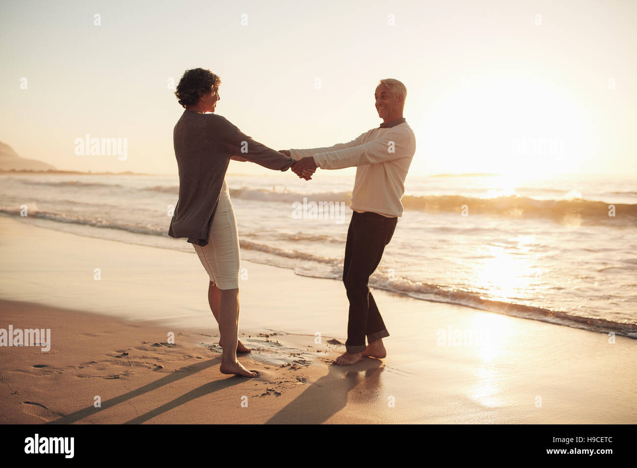Lunghezza piena ripresa esterna della coppia senior tenendo le mani in piedi sulla spiaggia. Uomo maturo e la donna giocando sulla spiaggia al tramonto. Foto Stock