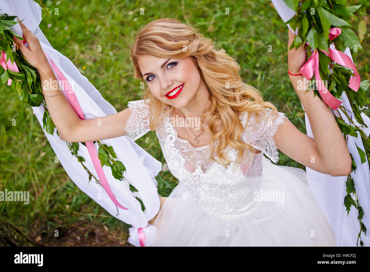 Bella bionda sposa in un bianco abito da sposa oscilla su decorate swing e sorridente per la fotocamera Foto Stock