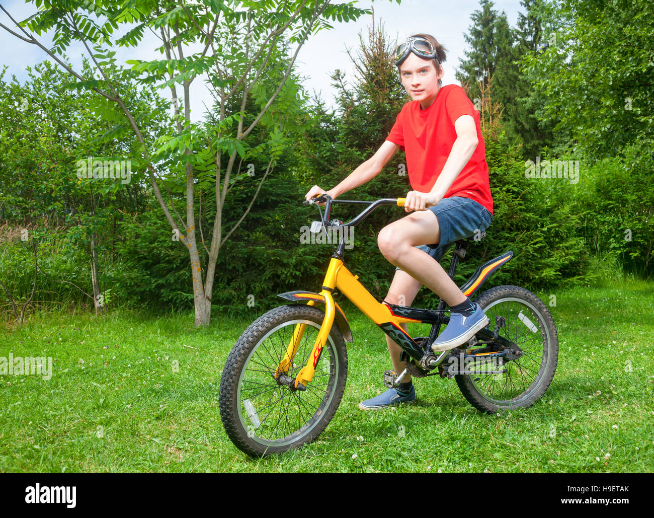 Carino teen ragazzo indossa tshirt rosso e gli occhiali seduto su una bicicletta in un parco d'estate guardando la fotocamera Foto Stock