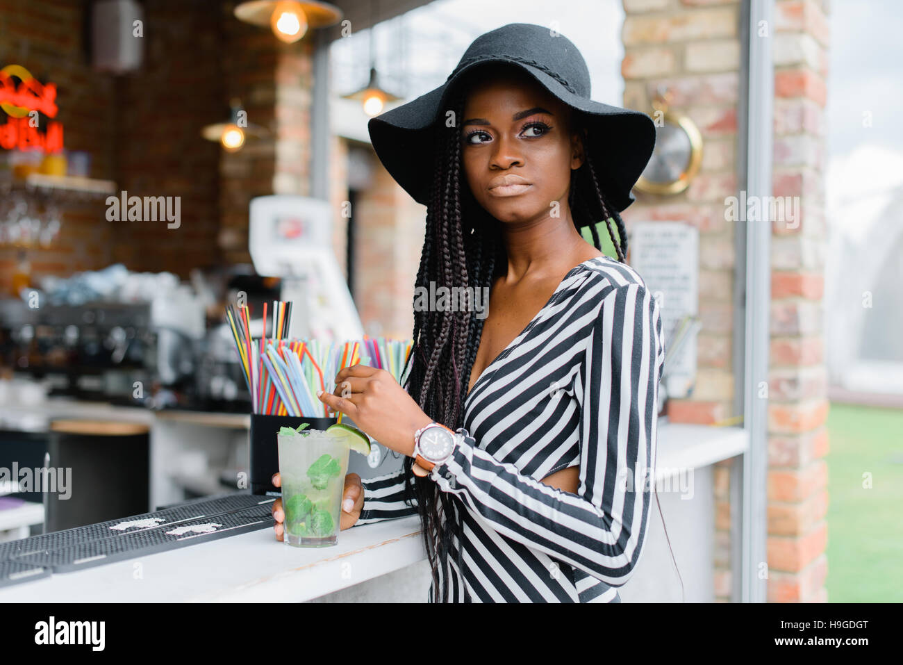 Attraente ragazza nera in bar con cocktail nelle sue mani. Molti cannucce per cocktail sul bar. Cocktail di ghiaccio. Mohito. Foto Stock