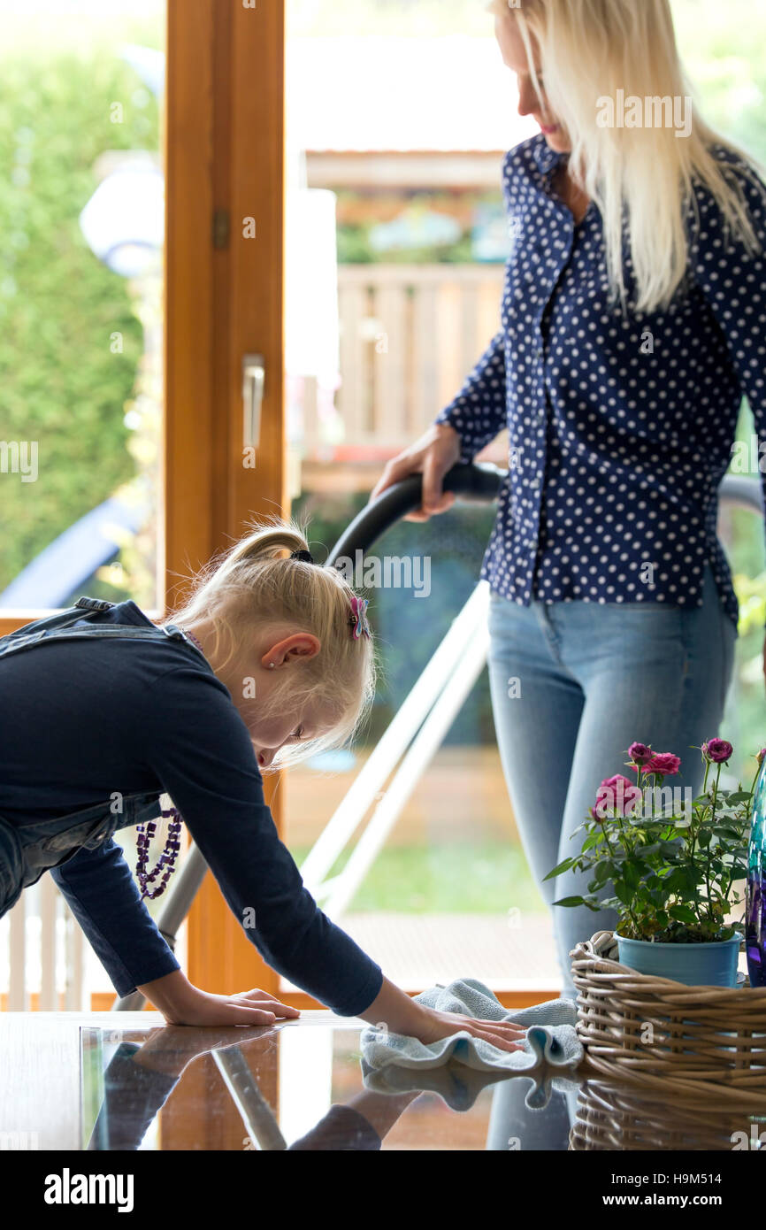 Ragazza pulizia vetro tavolo mentre sua madre hoovering Foto Stock