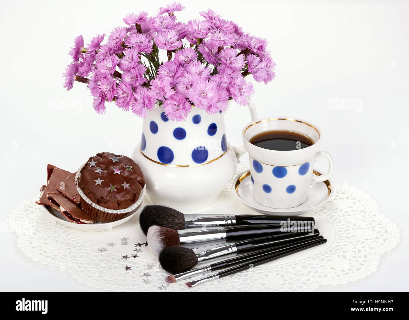 Pennelli per il trucco, fiori nella brocca e tazza di caffè con dei pirottini su sfondo bianco Foto Stock