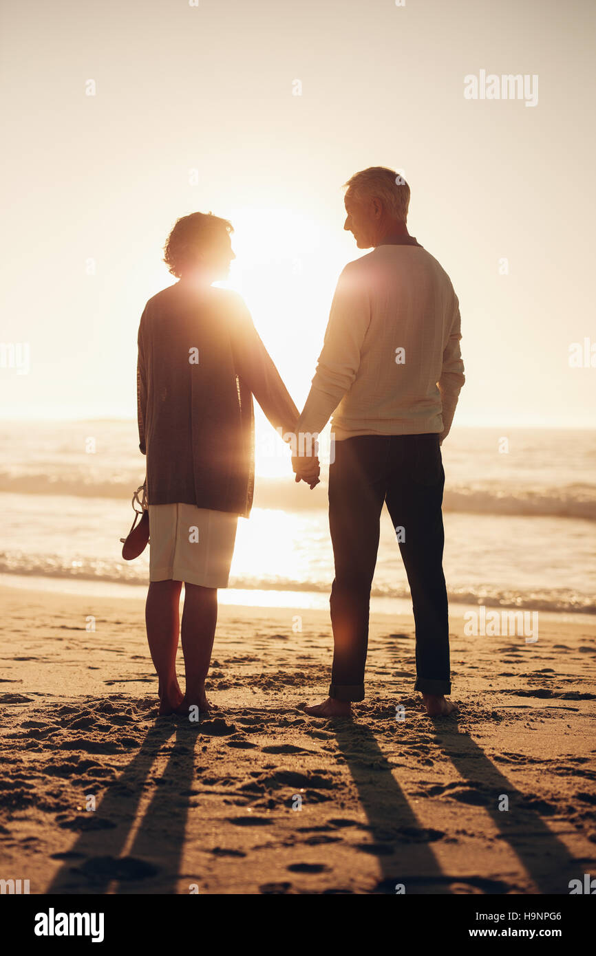 Vista posteriore del colpo di un amorevole coppia senior in piedi con la mano e guardando ogni altro sulla spiaggia. Coppia matura sulla riva del mare al tramonto. Foto Stock