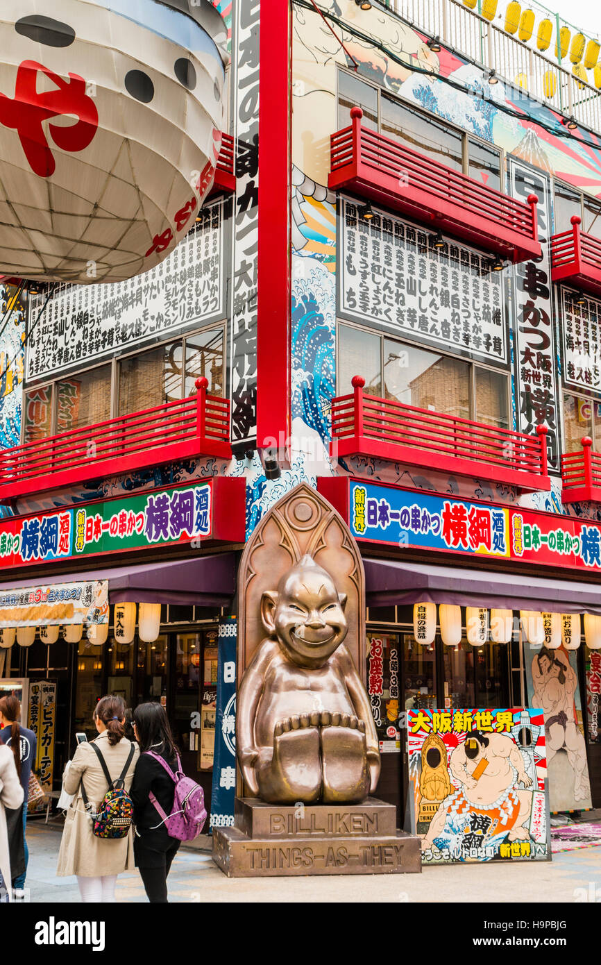 Giappone, Osaka, Shinsekai. Famosa statua Billiken, mascotte locale al di fuori su un angolo del ristorante Osaka vendita specialità alimentari, Kushi-katsu. Foto Stock