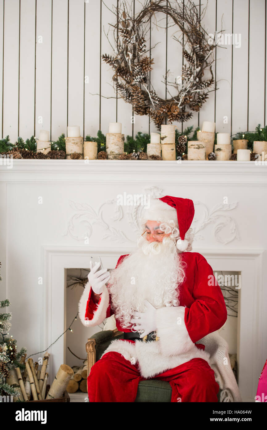 Felice di Santa Claus la digitazione di un messaggio sul telefono cellulare e sorridenti mentre è seduto alla sua sedia con camino. Albero di Natale in background Foto Stock