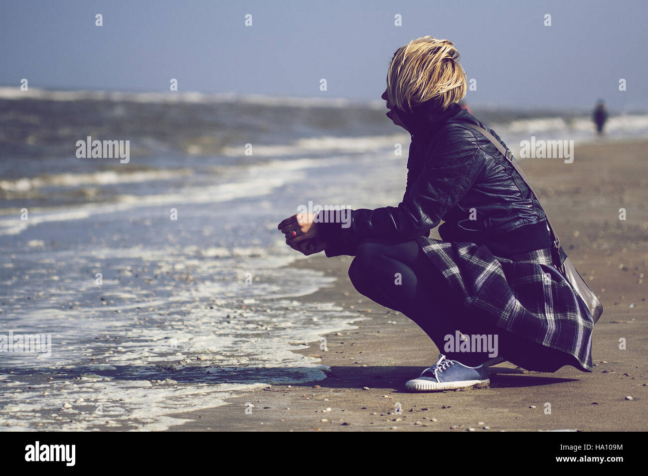 Giovani caucasici donna bionda con pelle nera giacca e gonna rannicchiato sulla sabbia di fronte al mare Foto Stock