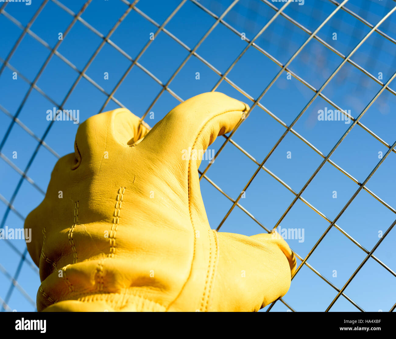 Pelle gialla maschio guantata mano afferra il filo filo di maglia contro un cielo blu sullo sfondo. Copyspace area per la costruzione e i concetti di allevamento. Foto Stock