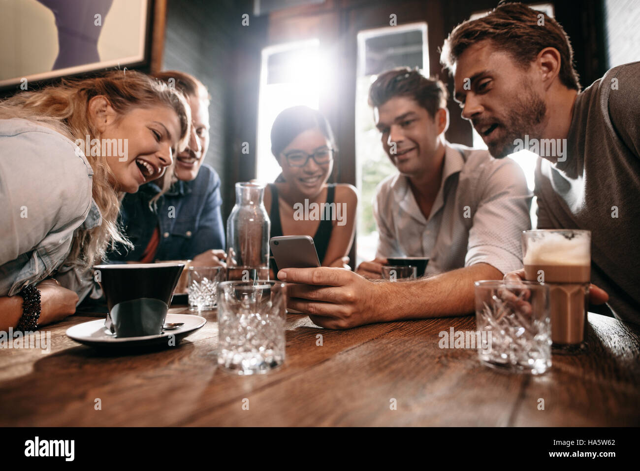 Giovani amici guardando smart phone mentre è seduto al cafe'. Razza mista persone sedute a un tavolo nel ristorante utilizzando il telefono cellulare. Foto Stock