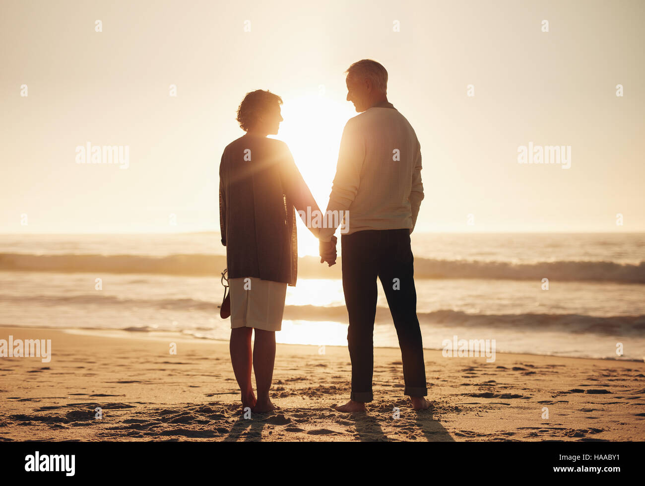 Vista posteriore di una coppia senior tenendo le mani sulla spiaggia. Coppia matura in piedi insieme su una spiaggia al tramonto. Foto Stock