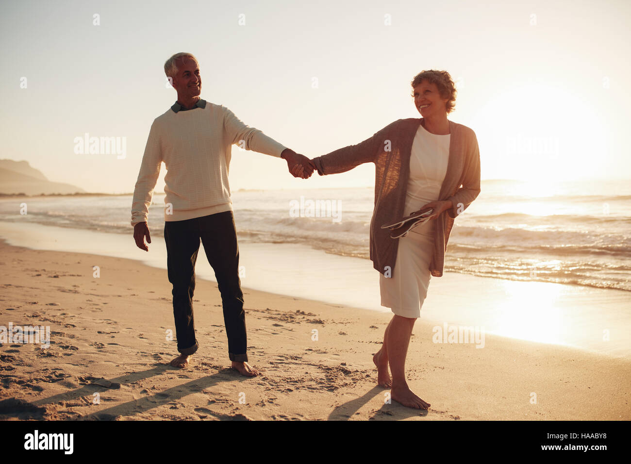 Lunghezza piena ripresa esterna di amorevole coppia matura camminando lungo la spiaggia tenendo le mani. Senior l uomo e la donna che cammina sulla riva del mare al tramonto. Foto Stock