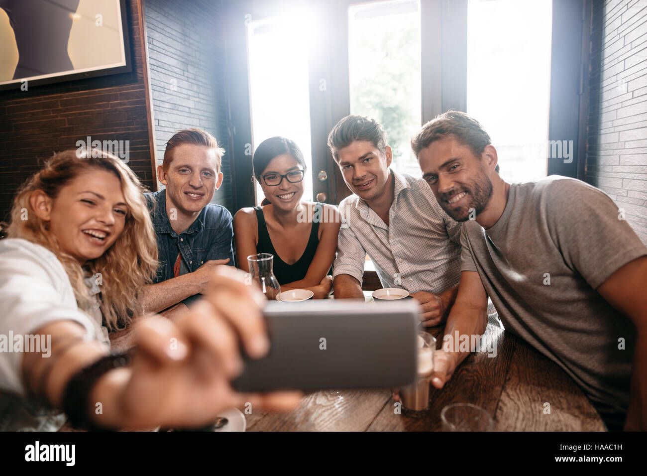 Piccolo gruppo di amici prendendo selfie su un telefono cellulare. Giovani uomini e donne seduti insieme al caffè e prendendo un autoritratto su smart phone. Foto Stock