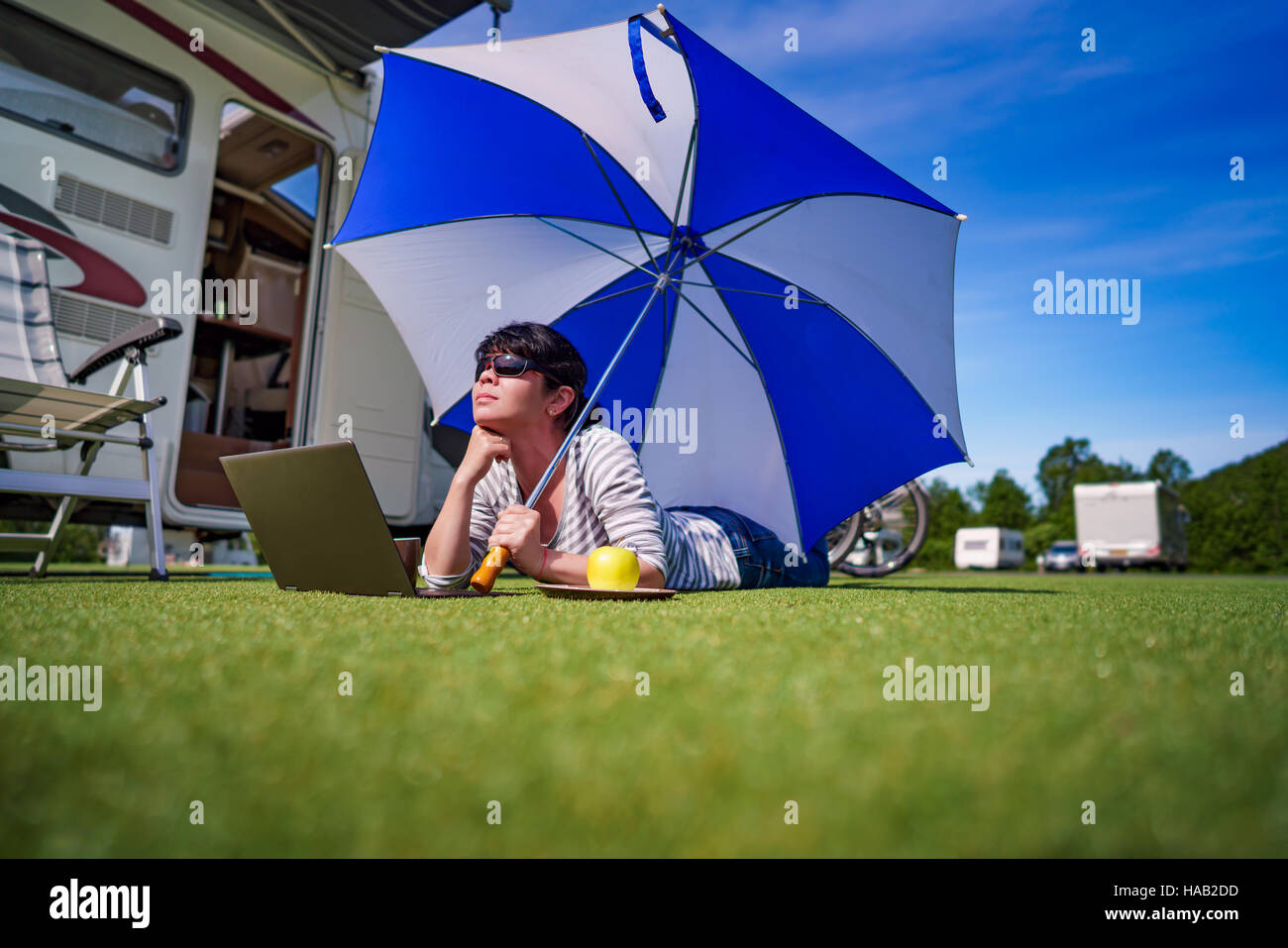 Donna sull'erba, guardando il laptop sotto ombrellone vicino al campe. Caravan auto vacanza. Vacanza per la famiglia in viaggio, viaggio vacanza in camper Foto Stock