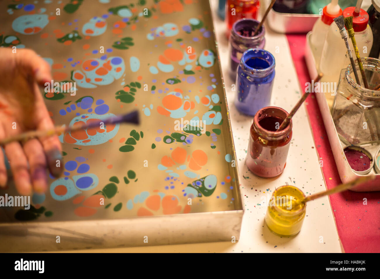 Ebru arte o marmorizzazione arte creazione nel processo. Donna mano spargendo vernice su acqua e vernice colorata bottiglie in piedi accanto al vassoio di ebru Foto Stock