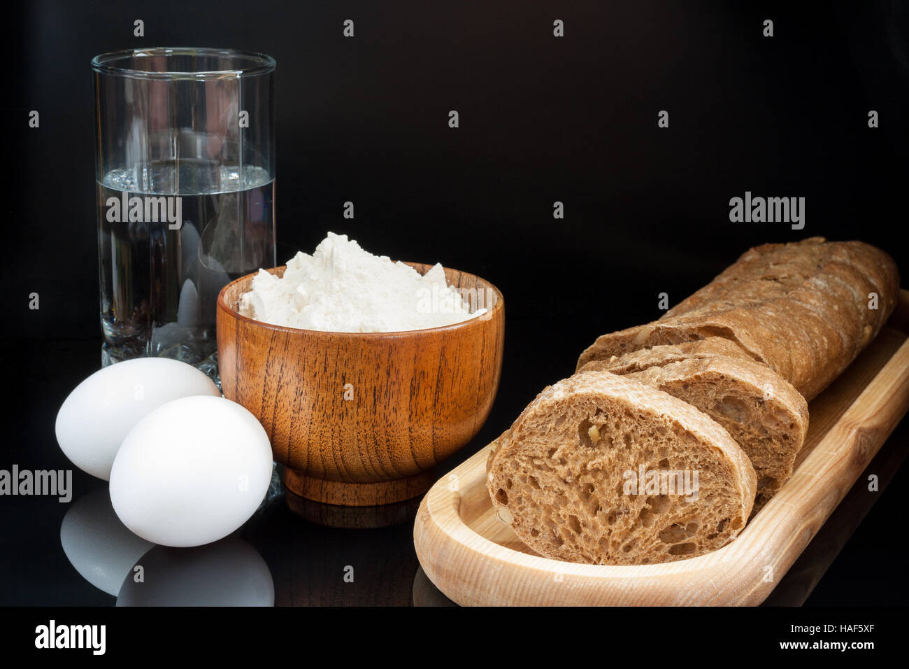 Ingredienti per la cottura del pane: farina, uova, bicchiere di acqua con  fresca baguette affettata su sfondo nero Foto stock - Alamy