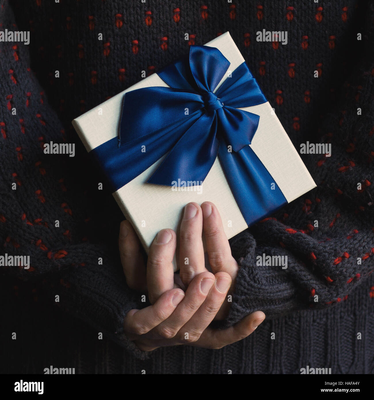 Close up di un uomo in maglia maglione scuro tenendo un bianco presente confezione regalo con nastro blu dietro la schiena Foto Stock