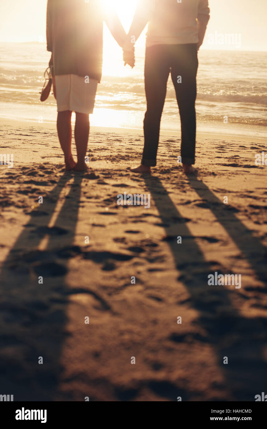 Vista posteriore mozzato colpo di una coppia senior in piedi con la mano sulla spiaggia. Coppia matura insieme sulla riva del mare al tramonto. Foto Stock