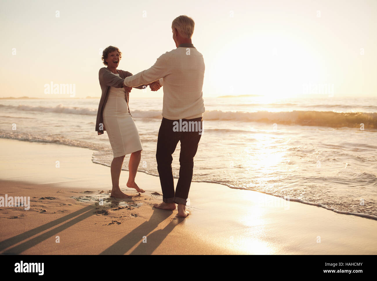 A piena lunghezza shot di sorridere coppia senior avendo divertimento sulla spiaggia. Giovane giocando sulla riva del mare tenendo le mani al tramonto. Foto Stock