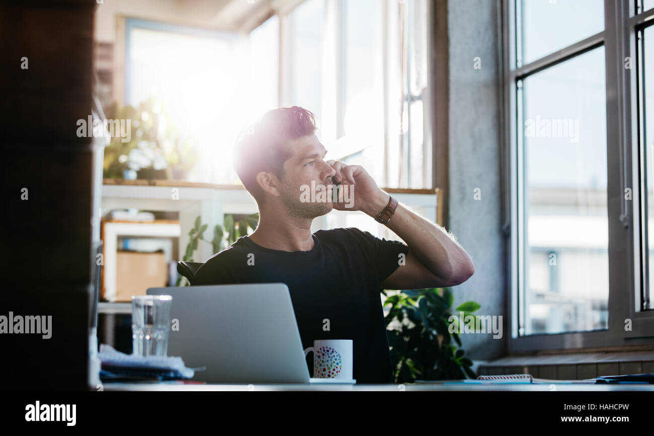 Colpo di giovane uomo seduto al suo ufficio parlando al cellulare. Business man lavorando alla sua scrivania con computer portatile effettuando una chiamata telefonica. Foto Stock