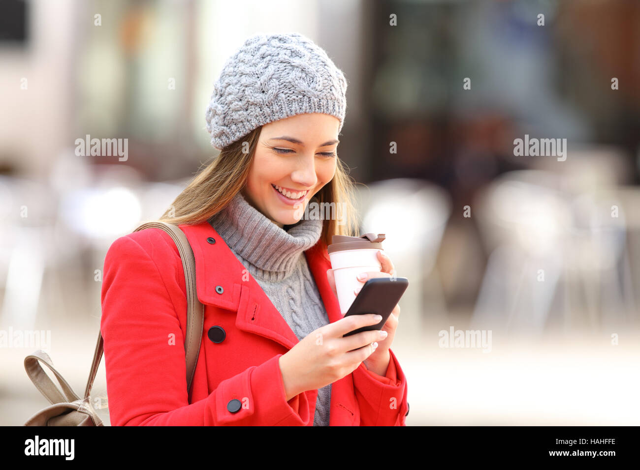Felice moda donna utilizzando uno smart phone e tenendo un take away caffè in strada in inverno Foto Stock