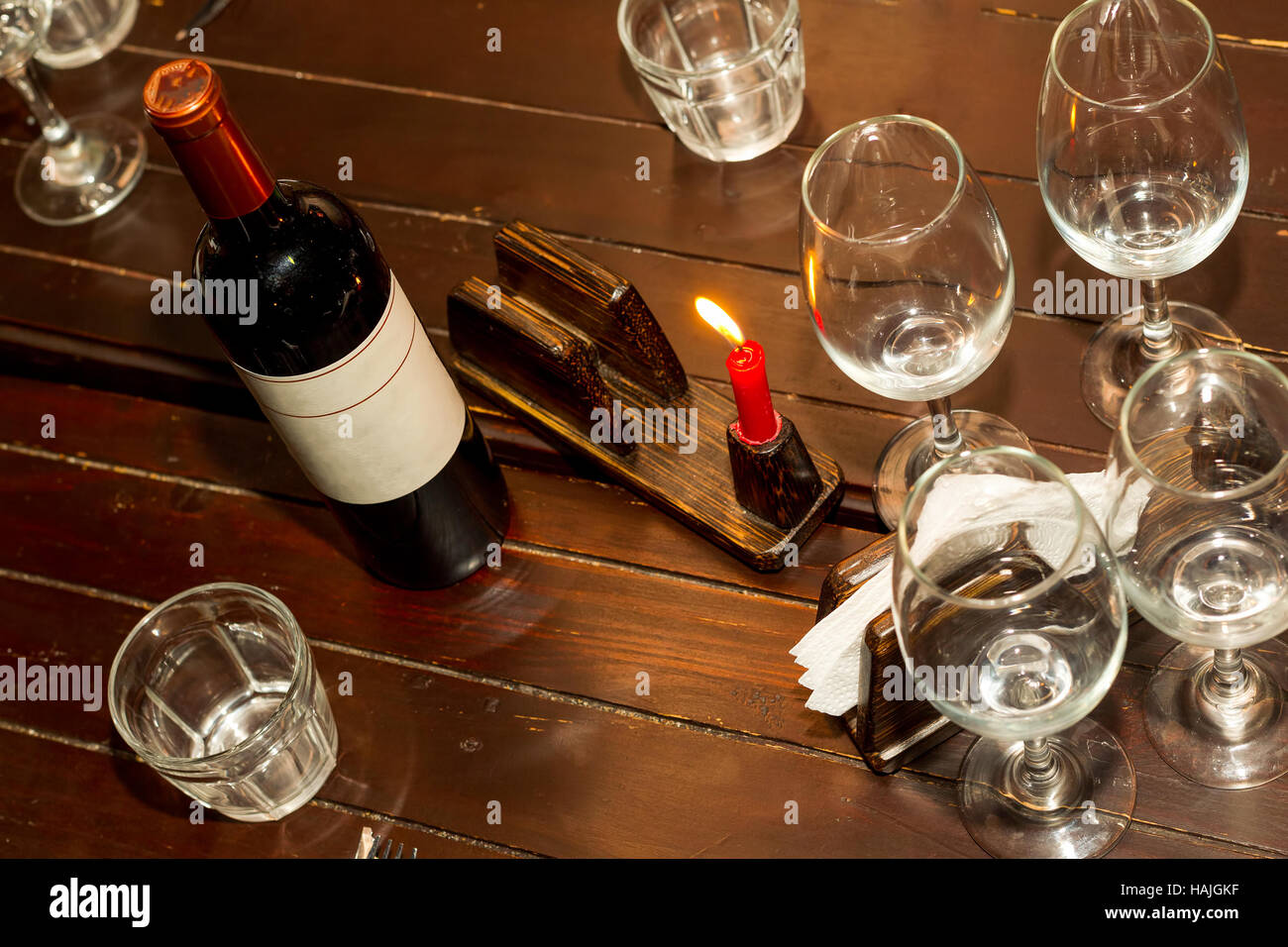 Bottiglia di vino rosso con molti bicchieri di vino vuote, seduto su un tavolo di legno illuminato da un lume di candela rossa Foto Stock