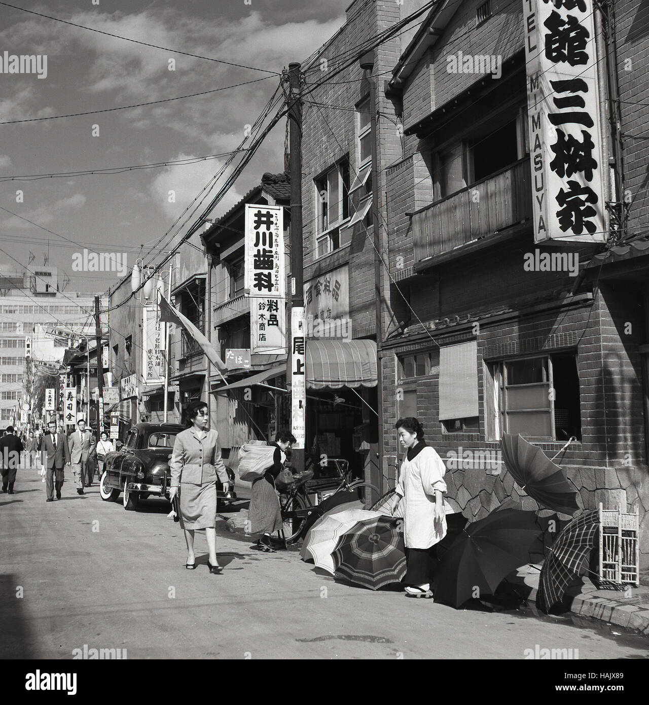 Anni '1950, storica, una donna commerciante di strada giapponese fuori da un edificio nella città vecchia che vende ombrelli, Tokyo, Giappone. Le piogge sono abbondanti nella città captali, in particolare nei mesi di settembre e ottobre, quando si verificano i tifoni. Foto Stock