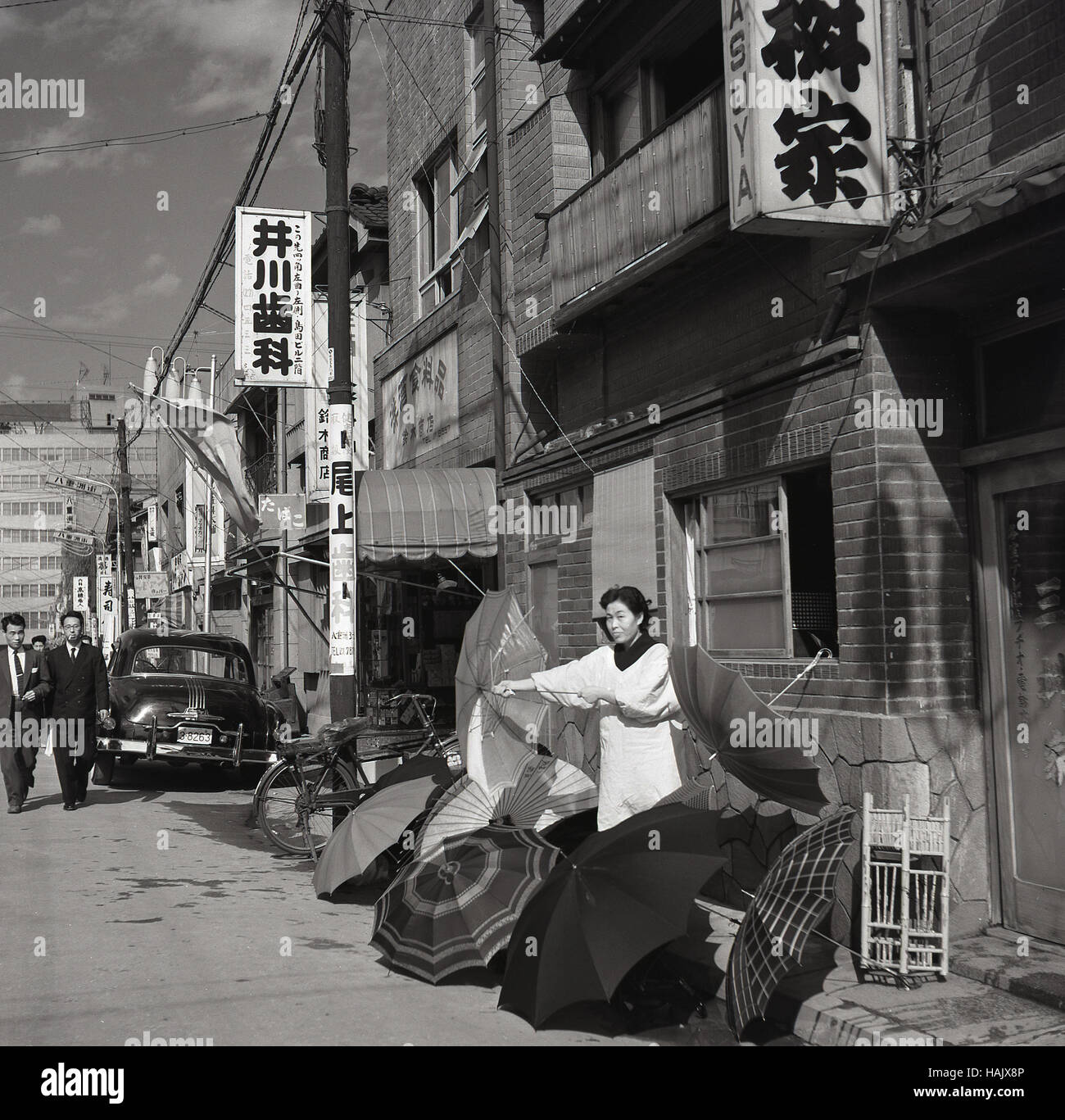 Anni '1950, storica, una donna commerciante di strada giapponese fuori da un edificio nella città vecchia che vende ombrelli, Tokyo, Giappone. La città captile è un luogo in cui le piogge sono abbondanti, in particolare in settembre e ottobre, quando si verificano i tifoni. Foto Stock