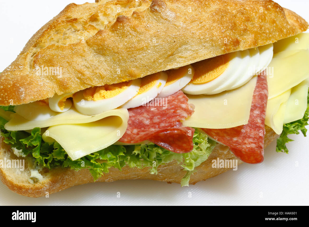 Baguette bun e rabboccato con salame, formaggio, lattuga e uova sode su un bianco tagliere in plastica Foto Stock
