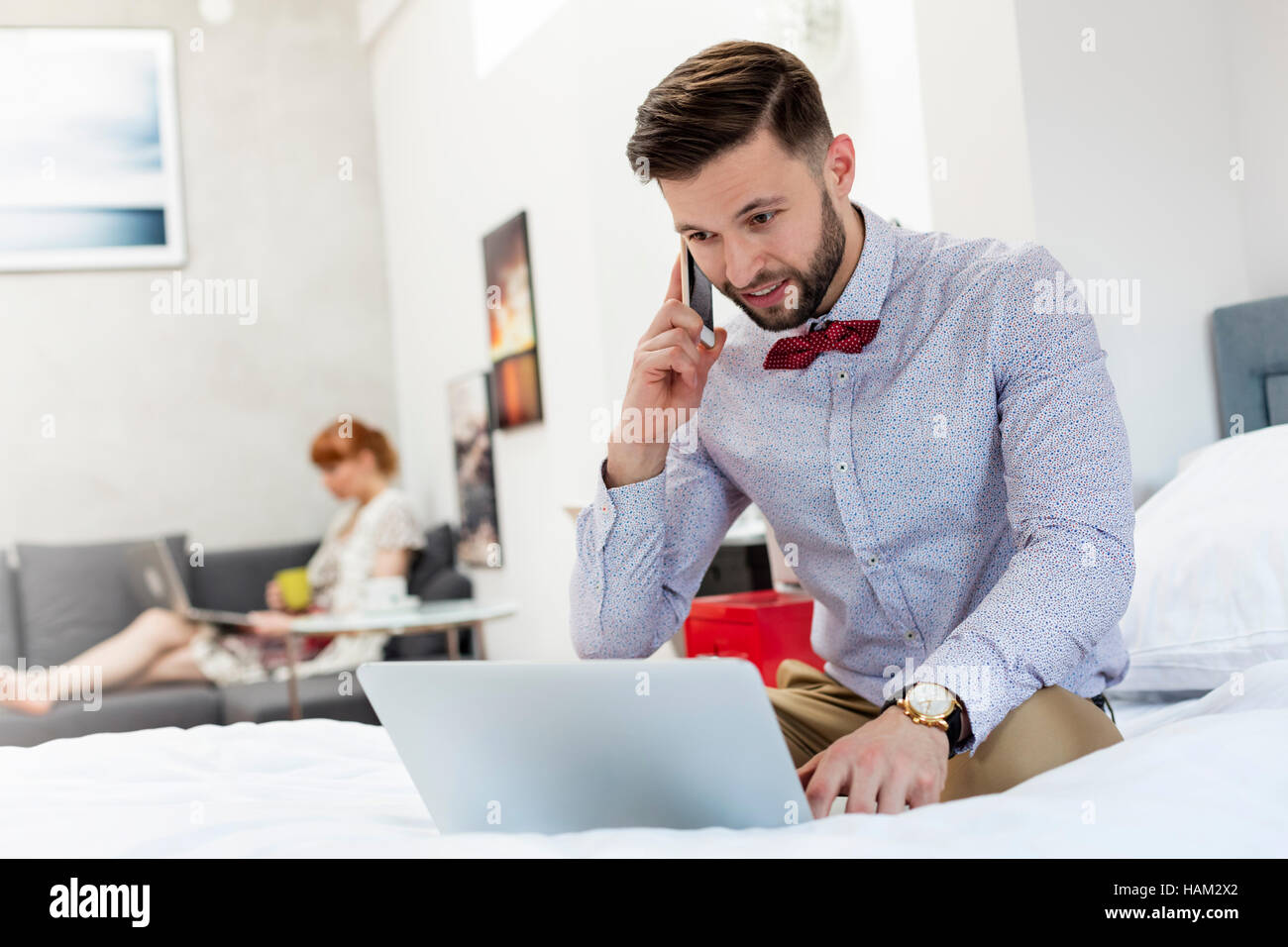 Uomo a bow tie parlando al cellulare e utilizzando computer portatile sul letto Foto Stock
