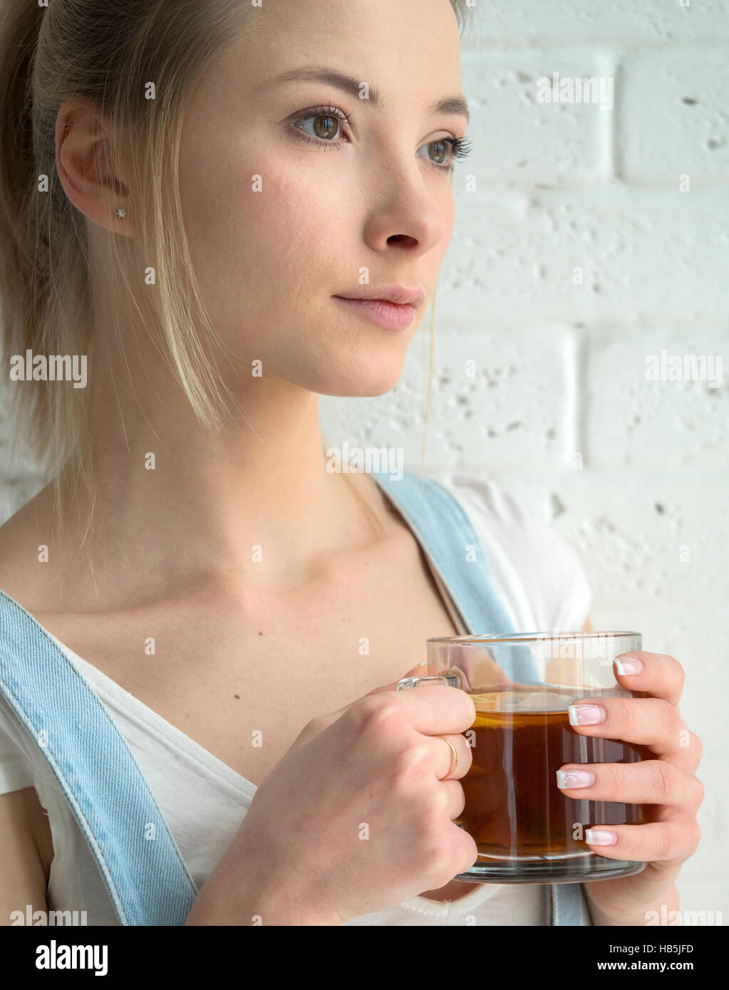 Bellezza giovane donna caucasica in denim blu con tuta e una T-shirt bianco bere il tè davanti un muro di mattoni Foto Stock