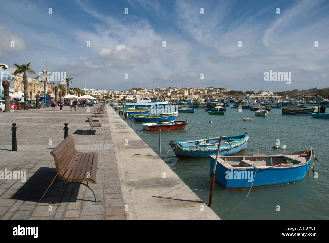 Vista sul porto del piccolo porto di pescatori di Marsaxlokk a Malta, con passeggiata, edifici e piccole barche da pesca contro un cielo blu Foto Stock