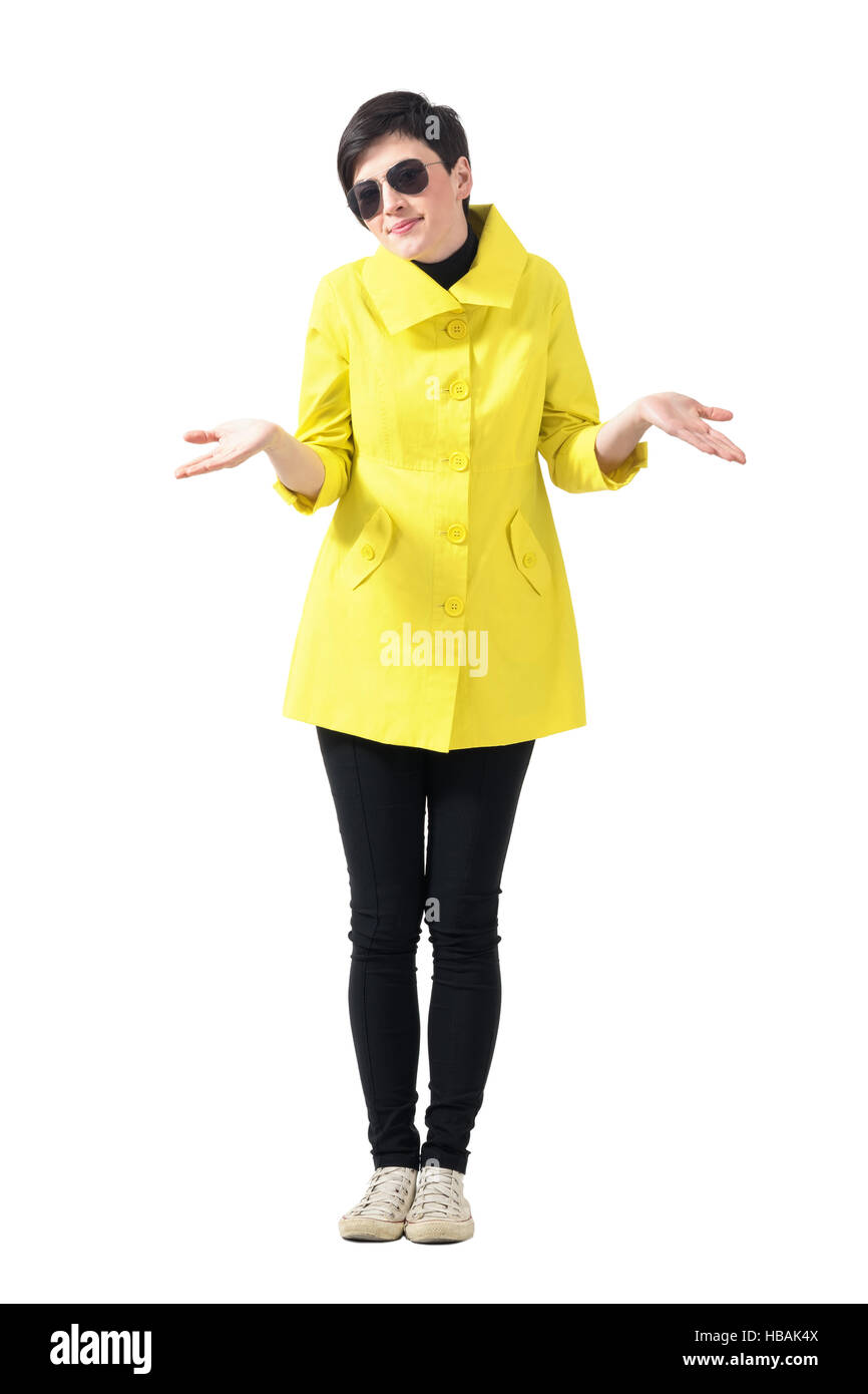 Confuso donna Cappotto giallo indossando occhiali da sole scrollare le spalle. Corpo pieno lunghezza ritratto isolate su sfondo bianco Foto stock Alamy