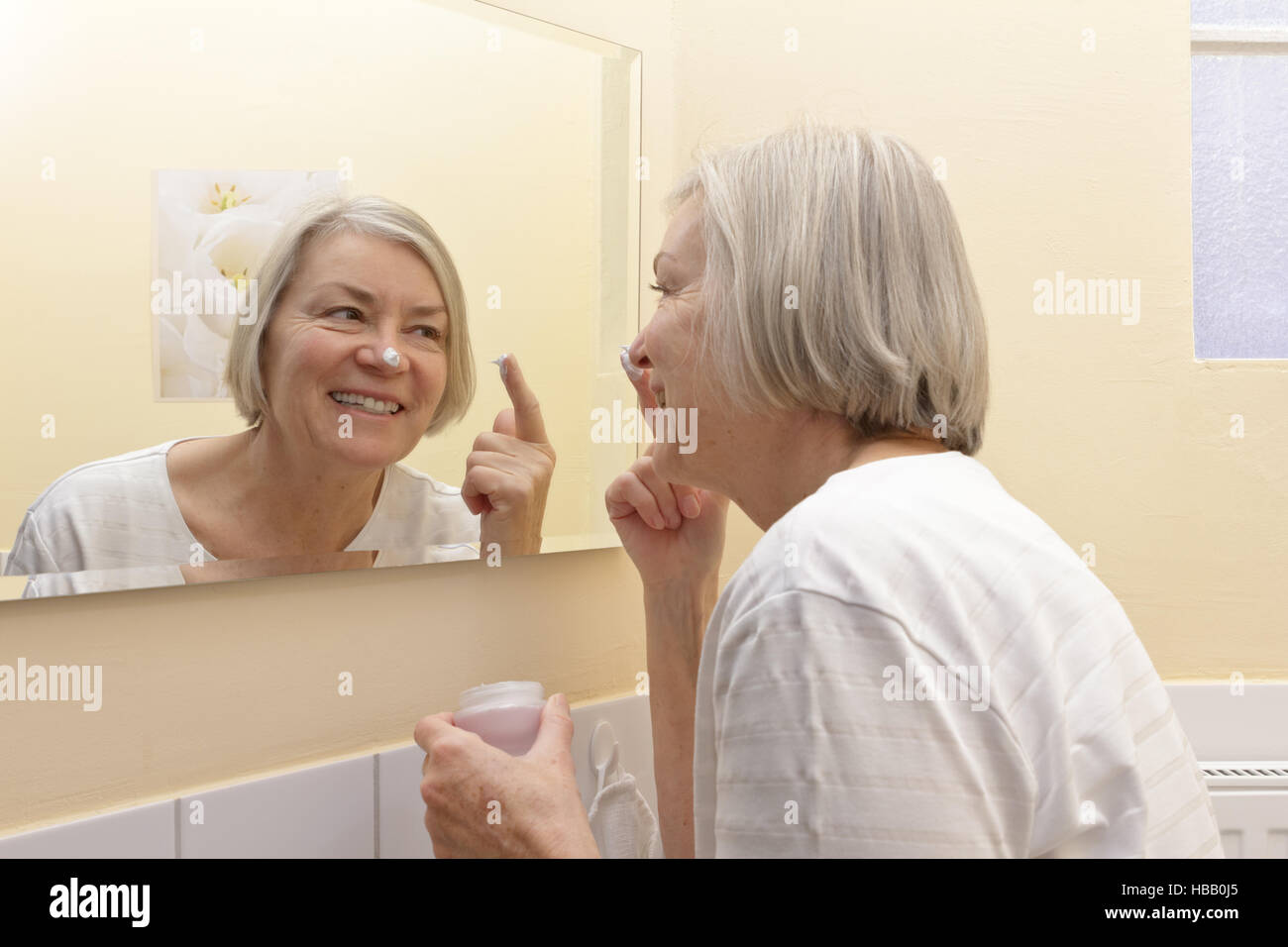 Vecchia donna con un grande sorriso e una macchia di crema sul naso di fronte ad uno specchio per applicare un idratante sul suo viso Foto Stock Vecchia donna con un grande sorriso e una macchia di crema sul naso di fronte ad uno specchio per applicare un idratante sul suo viso Foto Stock