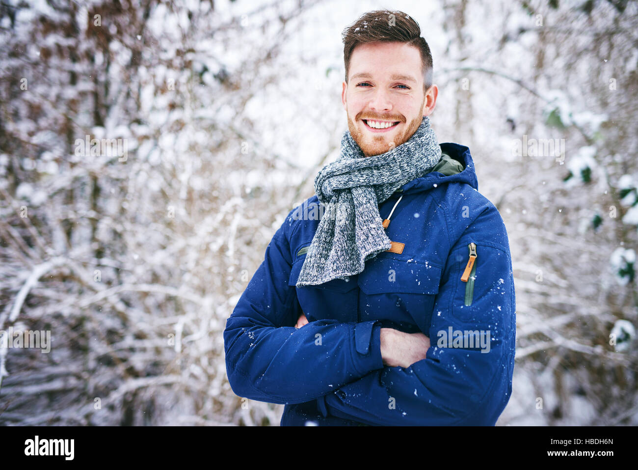 Giovane uomo in cappotto in posa con mani incrociate e sorridente alla fotocamera su sfondo di boschi innevati. Foto Stock