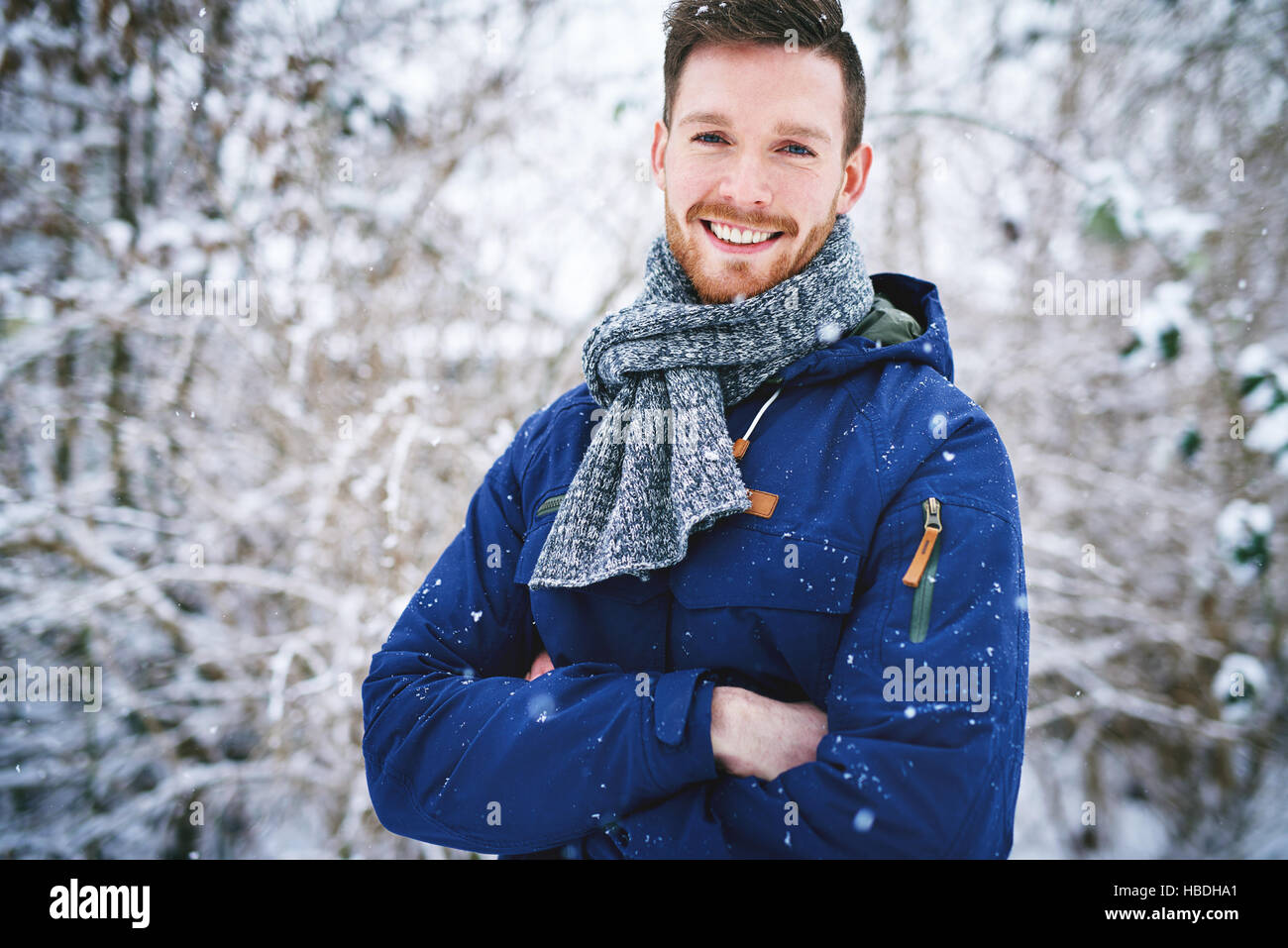 Giovane uomo bello in giacca calda con mani incrociate sorridente alla fotocamera su sfocata foreste innevate sullo sfondo. Foto Stock