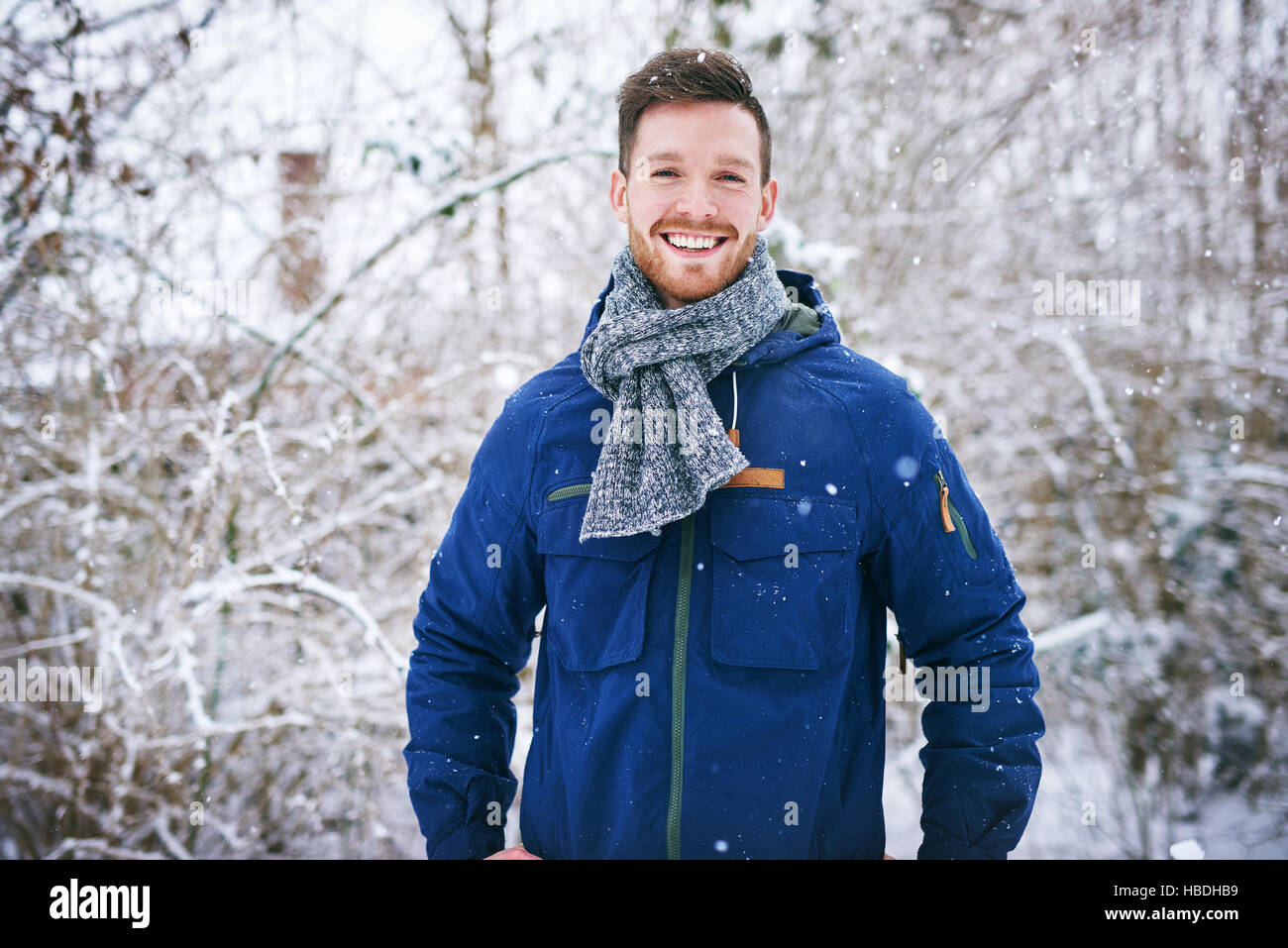 Giovane uomo barbuto uomo in caldo cappotto sorridente alla fotocamera su sfondo sfocato dei boschi innevati. Foto Stock