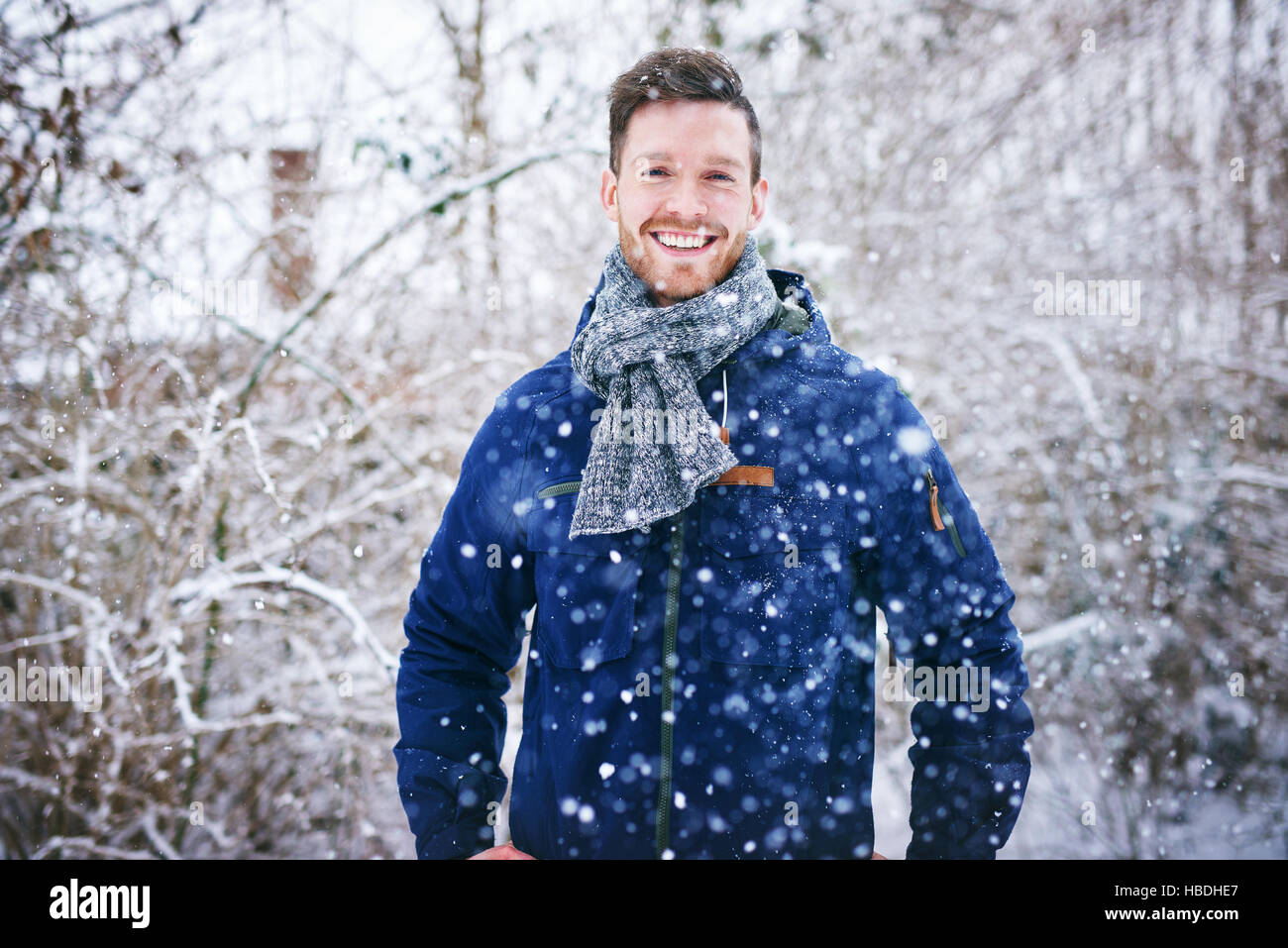 Giovane uomo bello in giacca calda sorridente in telecamera in piedi nella neve sullo sfondo dei boschi innevati. Foto Stock
