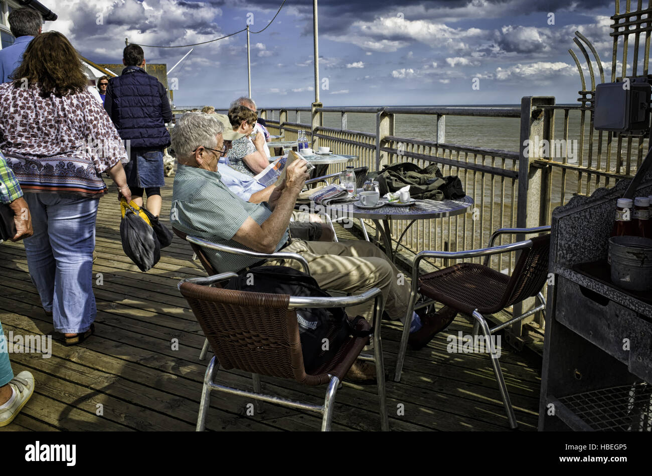 Southwold, England Regno Unito. Le persone anziane godendo il sole seduti lungo Southwold pier bere il tè e la lettura dei quotidiani. I visitatori più giovani sono a piedi da. Fotografia convertito in HDR, High Dynamic Range , per un più drammatico alto contrasto dell'immagine. Foto Stock