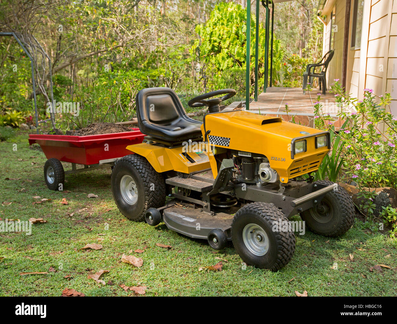 Giallo ride-sulla falciatrice piccola di traino rimorchio rosso pieno di suolo sul prato accanto a casa con giardino lussureggiante di arbusti e alberi Foto Stock