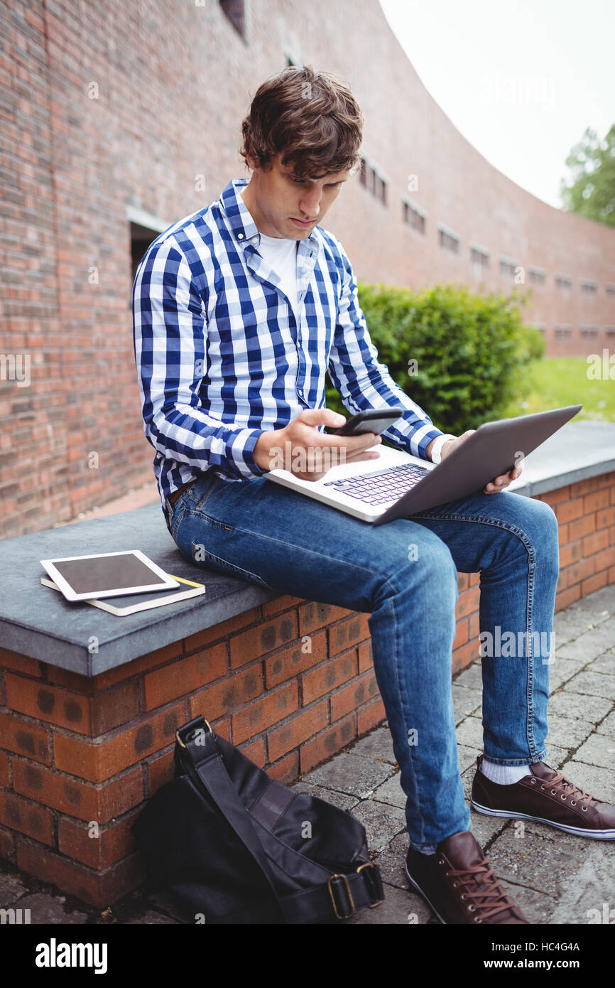 Studente seduto con il computer portatile utilizzando il telefono cellulare nel campus Foto Stock