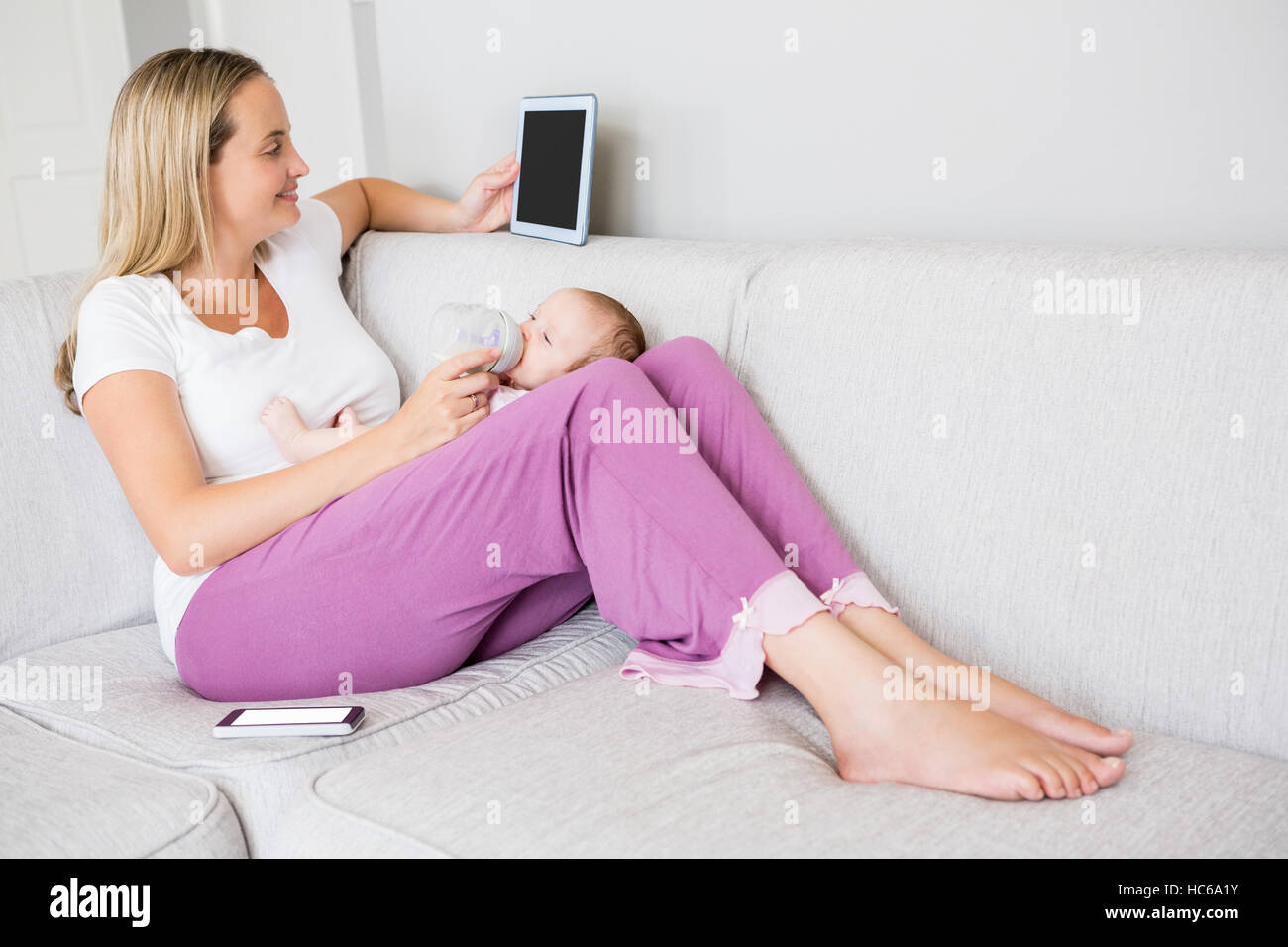 Madre utilizzando tavoletta digitale mentre si alimenta il suo bambino con bottiglia di latte Foto Stock